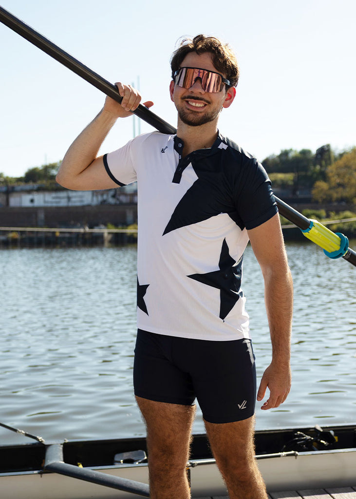 Man wearing a black and white star-patterned henley and black trou, holding an oar on a lake.