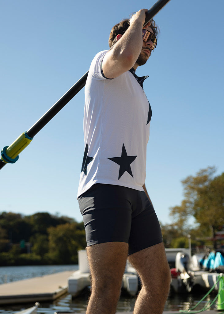 Person standing with an oar with a star design on their shirt, standing on a dock with trees and water in the background.